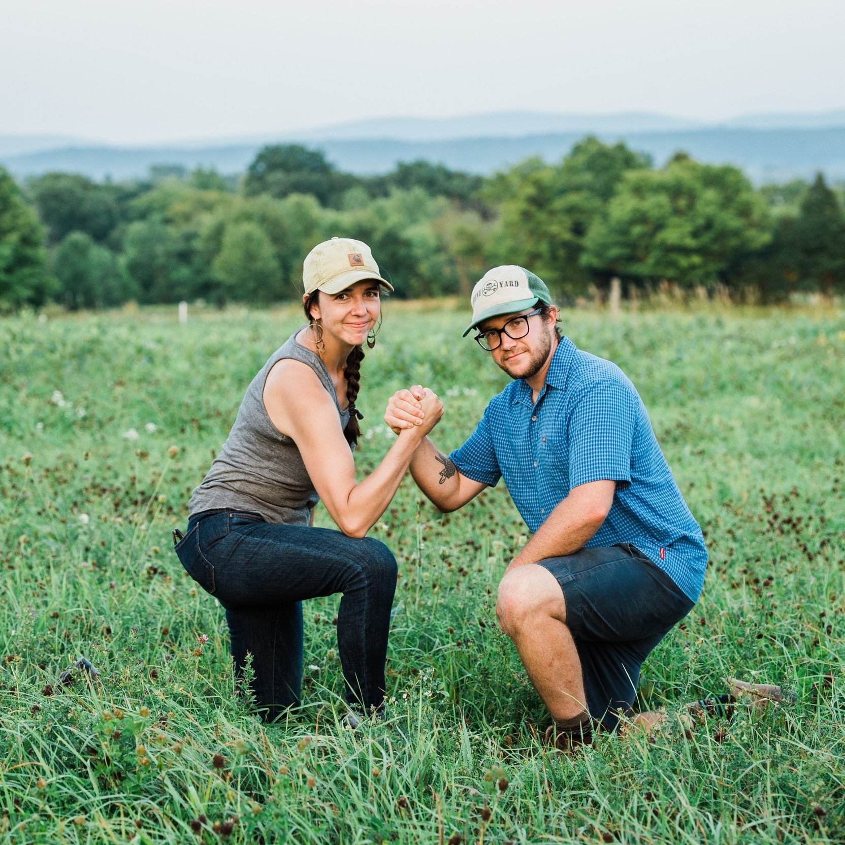 A woman and a man engaged in a friendly arm wrestling match outdoors on a farm, surrounded by green grass and trees, showcasing a playful atmosphere in a rural setting.